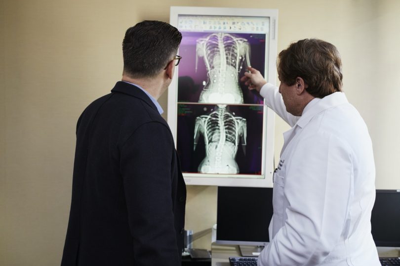 doctor pointing x ray result beside man wearing black suit 810x540